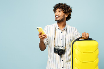 Traveler Indian man wear white casual clothes hold bag use mobile cell phone isolated on plain blue background. Tourist travel abroad in free spare time rest getaway. Air flight trip journey concept.