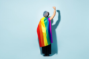 Fototapeta premium Full body back rear view young happy gay man he wrapped in striped rainbow flag clenching fist isolated on plain pastel blue wall background studio portrait. Pride day June month love LGBT concept.