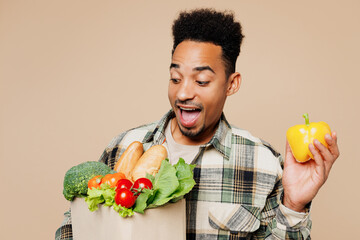 Young man wear grey shirt hold yellow bell pepper look at paper bag for takeaway mock up with food products isolated on plain pastel light beige background. Delivery service from shop or restaurant.