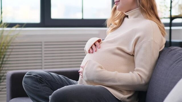Close up shot of a pregnant Caucasian young woman stroking belly, relaxing on sofa at home living room. Pregnancy, childbirth, future parenthood concept.