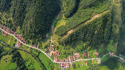 Drone panorama above a village located inside a mountainous area, along a river. The river and a road winds near beech forests, and meadows. Countryside landscape, Carpathia, Romania.