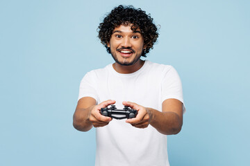 Young fun happy Indian man he wear white t-shirt casual clothes hold in hand play pc game with joystick console isolated on plain pastel light blue cyan background studio portrait. Lifestyle concept.