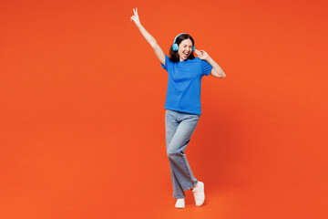 Full body young smiling cool happy woman she wears blue t-shirt casual clothes listen to music in headphones raise up hand isolated on plain red orange background studio portrait. Lifestyle concept. - obrazy, fototapety, plakaty