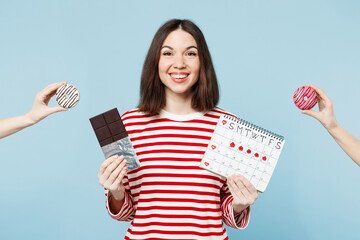 Young smiling happy woman wear red casual clothes eat chocolate bar donuts hold female periods pms calendar checking menstruation days isolated on plain blue background. Medical gynecological concept.