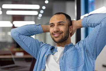 Close-up photo of satisfied and smiling young Arab man in casual clothes sitting in modern office...