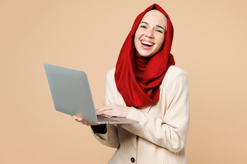 Young Arabian Asian Muslim IT woman wear red abaya hijab suit clothes hold use work on laptop pc computer laugh isolated on plain beige background studio. UAE middle eastern Islam religious concept