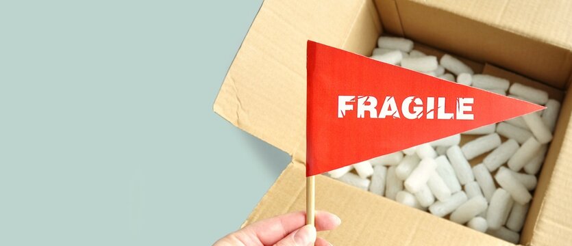 Opened cardboard packaging box with white polystyrene packing chips inside. Tiny red paper flag with the warning 'Fragile' as a label, sticker. The concept of packing and shipping fragile items