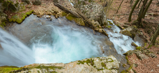 An azure spring flowing downhill across mossy, sharp rocks. The rapid stream forms numerous...