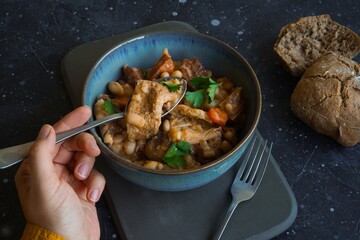 A female hand with spoon of Tripas - pork tripe stew with white beans and smoked sausages in a bowl on the dark table