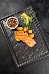 Salmon steak with baked potatoes and sauce on black stone plate. Salmon fillet roasted on grill with vegetable garnish in minimal style on dark background. Grilled salmon with potato in menu. - 788219201