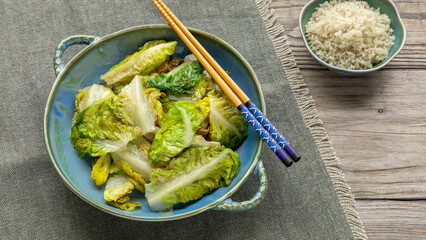 Stir-fried lettuce in a bowl. Prosperity dish served around Chinese New Year. Grey sea salt in a small bowl. Chopsticks on a bowl with lettuce and napkin on a wooden table. 