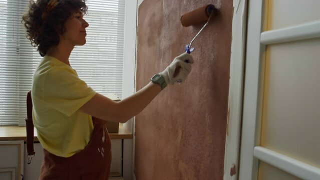 Side footage of positive Caucasian woman in late pregnancy stage painting walls with brown color using paint roller while remodeling room into nursery for unborn baby