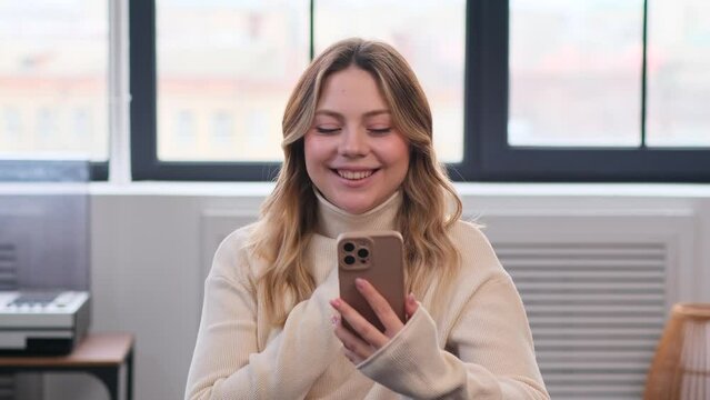 Happy Caucasian young woman laughing while relaxation with mobile phone on couch at home. Online messaging or internet browsing concept.
