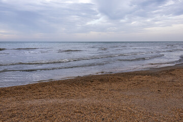A beach with small shells and the sea. The coast of the Caspian Sea. Cool weather on the coast. A walk along the seashore. The healing sea air. Waves on the surface of the water. Cloudy weather at sea