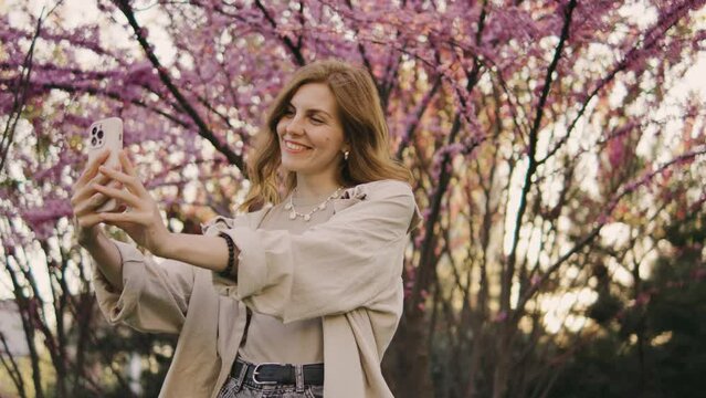 A happy young red-haired woman makes a selfie on the background of a tree that blooms with pink flowers. Selfie on a walk, blog, happy moments on camera. Spring flowers of cherry or sakura blossoms on