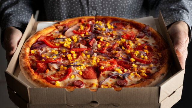 Caucasian male hands hold large open carton box with pepperoni pizza with sweetcorn front view