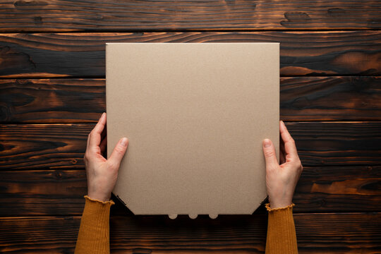 Pizza closed carton box in caucasian hands on natural dark wooden table flat lay mockup with copy space