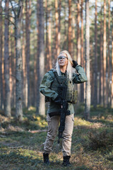Beautiful blonde military girl with a modern AK12 suppressed rifle in the forest at war. Vertical...