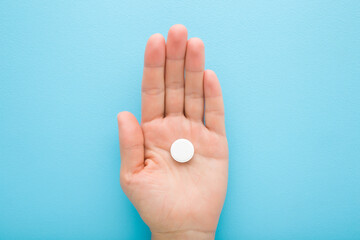Young adult woman opened palm holding white pill of c vitamin on light blue table background. Pastel color. Receiving nutrition supplement. Closeup. Top down view.