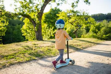 Adorable little boy riding his scooter in a city park on sunny summer evening. Young child riding a...
