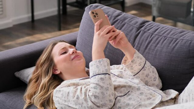 Friendly Caucasian young woman in pajama spending weekend leisure at home, relaxing on sofa with cellphone. Messaging in online social media.