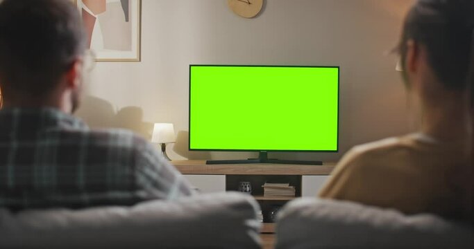 Rear view of married couple sitting at sofa. Young couple sitting in front of TV set with chroma key screen. Wife and husband looking at green mock up screen. Woman and man at living room.