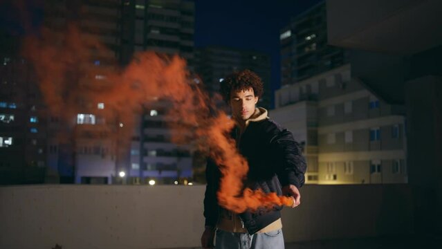 Hipster holding smoke bomb at night city. Curly guy teenager burning red grenade
