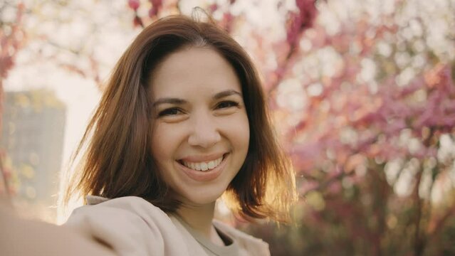 A happy young woman makes a pov selfie on the background of a tree that blooms with pink flowers. Selfie on a walk, blog, happy moments on camera. Spring flowers of cherry or sakura blossoms on