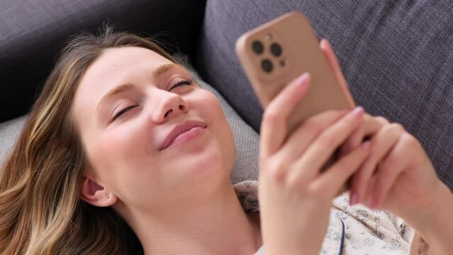 Close up portrait of a joyful Caucasian woman using phone and relaxing on sofa at home. Surfing internet, engaged in online entertainment.