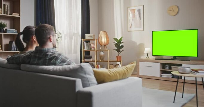 Couple sitting at sofa watching green chroma key screen. Woman and man sitting at couch looking at TV screen. Married couple relaxing at home watching chroma key TV set. Chroma key screen.