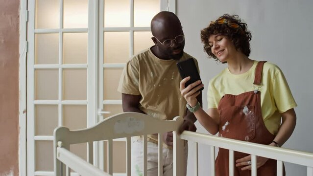 Medium long footage of Caucasian pregnant wife showing design ideas of nursery on mobile to African American husband while remodeling room for future offspring