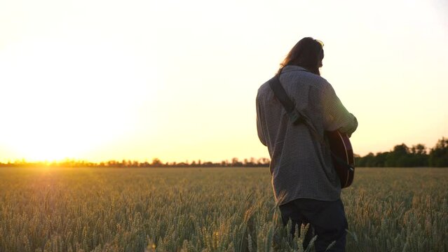 Male hipster playing on electric guitar at wheat field. Hippie man with long hair plays on musical instrument at barley meadow. Young guy resting at nature. Beautiful sunset at background. Slow motion