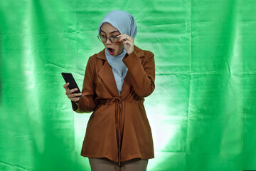 Shocked young Asian woman wearing hijab, glasses and blazer using mobile phone and taking of...