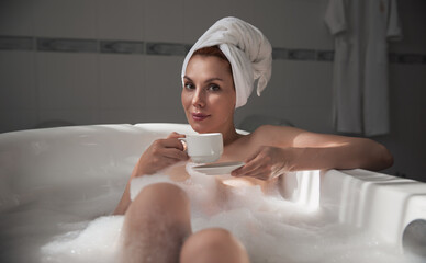 Relaxed serene young woman in spa bathroom, drinking tea, water. Stress Relief Concept. Woman with towel on her head, taking bath. Girl enjoying bath. Lady lying in bathtub. Girl relaxing in bathtub