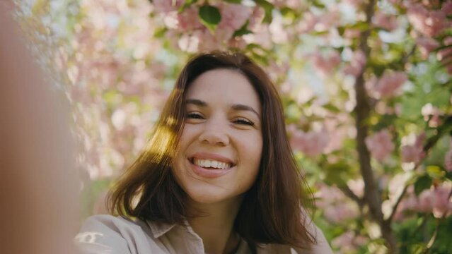 A happy young woman makes a pov selfie on the background of a tree that blooms with pink flowers. Selfie on a walk, blog, happy moments on camera. Spring flowers of cherry or sakura blossoms on