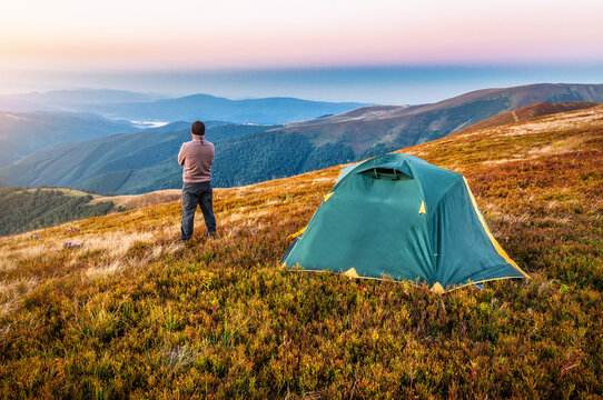Tourist hiker standing near tent and looking on mountains at sunset