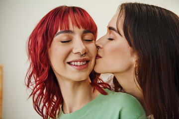 portrait of young and happy lesbian woman kissing cheek of her female partner with red hair at home