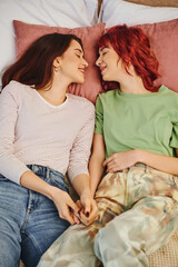 top view of smiling lesbian couple holding hands and lying together on bed, looking at each other