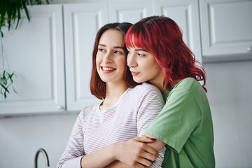 portrait of joyful and pierced lesbian woman with red hair embracing her girlfriend at home