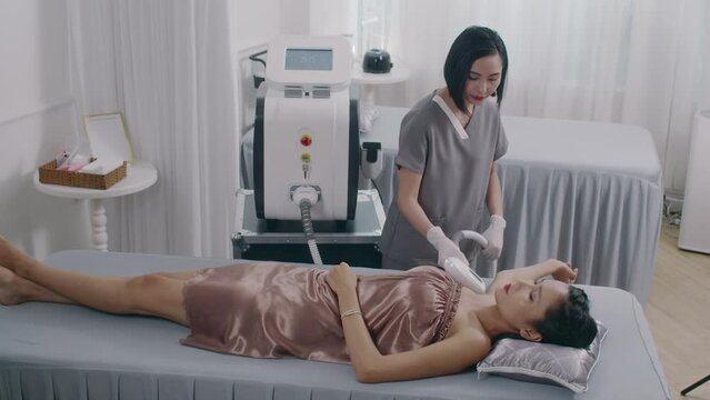 Wide shot of young Asian beautician doing armpit laser epilation to female client in spa salon