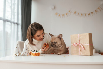 Hats, gift box and little cake for animal who have birthday party. Young woman is with her pug dog at home