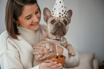 Celebration of birthday. In hats and with cake. Young woman is with her pug dog at home