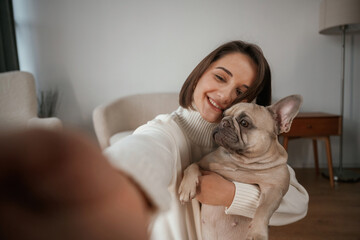 Making selfie, smiling. Young woman is with her pug dog at home