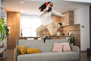 Woman with wireless headphones jumping on sofa and singing at home