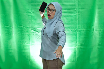 angry young Asian woman wearing hijab, glasses and blouse with displeased expression and wanna throw mobile phone over green background
