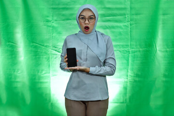 Shocked Asian woman wearing hijab, glasses and blouse showing blank smartphone screen with palm over green background
