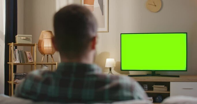 Man sitting at sofa at modern appartment. Male holding remote control and changing channel. Man looking at chroma key TV set. Male sitting at couch and looking at green moke up TV.