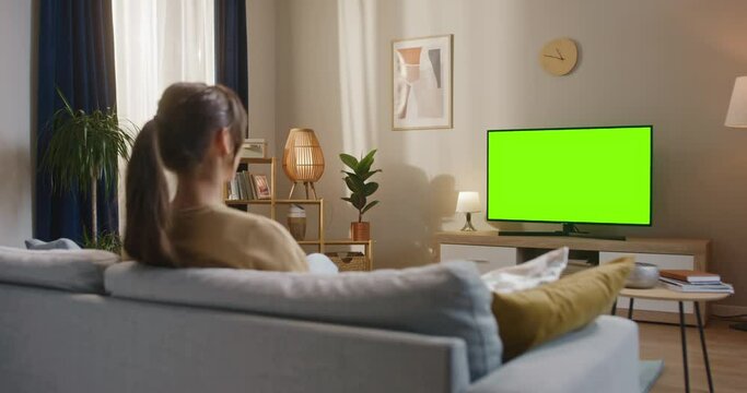 Woman sitting at sofa at modern appartment. Female holding remote control and changing channel. Female looking at chroma key TV set. Woman sitting at couch and looking at green moke up TV.