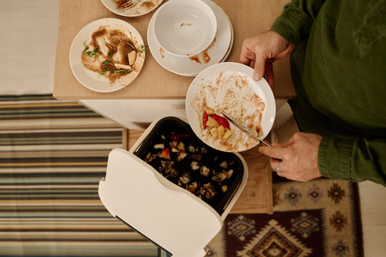 Top view of hands of young unrecognizable man with plate throwing meal leftovers in plastic bucket or trash bin containing soil