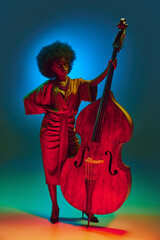 Elegant African-American woman with curly hairstyle in red dress poses with cello in neon light...